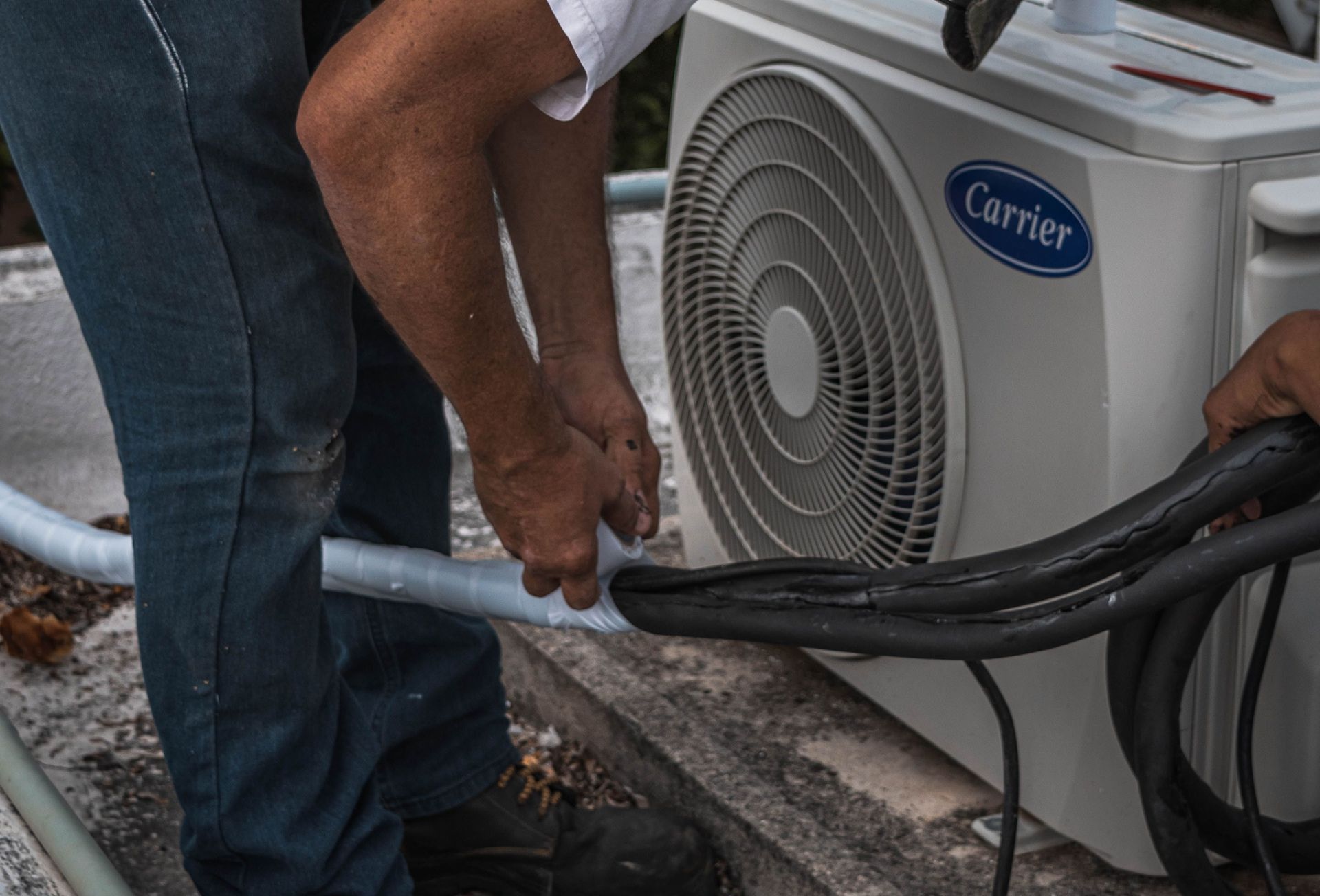 A technician performs maintenance on an outdoor air conditioning unit, focusing on hose connections.