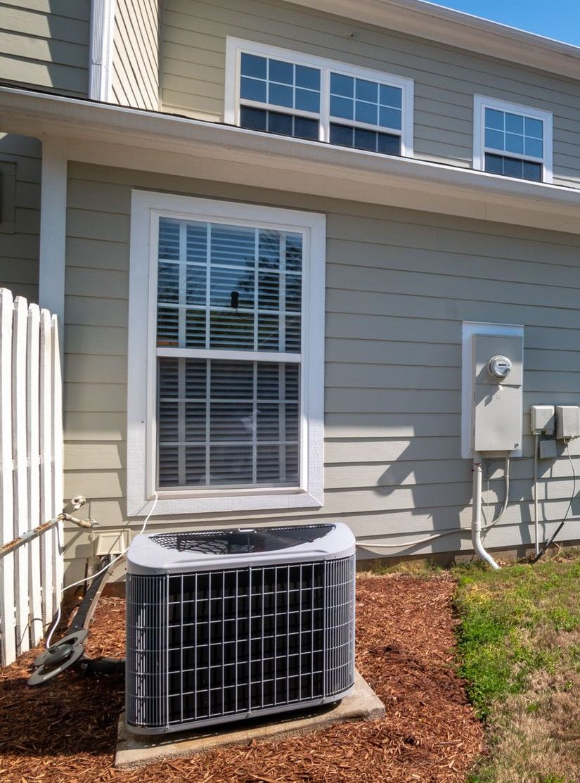 A sunlit backyard view of a modern grey house with white fencing and AC unit.